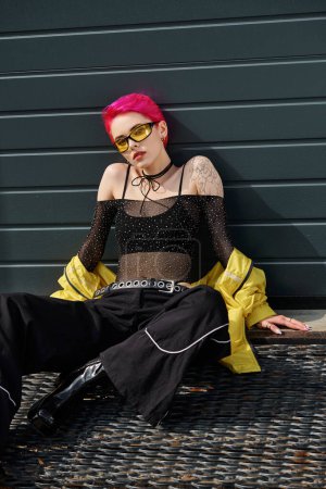 hübsche Frau mit pinkfarbenen Haaren und Tätowierung posiert in Sonnenbrille und trendiger Streetwear auf urbaner Straße
