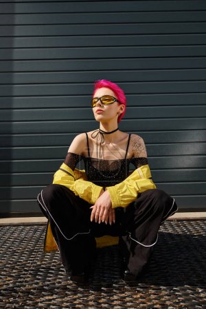 Charmante Frau mit pinkfarbenen Haaren und Tätowierung posiert in Sonnenbrille und trendiger Streetwear auf urbaner Straße