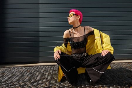 verführerische Frau mit pinkfarbenen Haaren und Tätowierung posiert in Sonnenbrille und trendiger Streetwear auf urbaner Straße