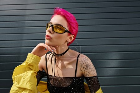 Foto de Mujer joven pensativa con pelo rosa y tatuaje posando en gafas de sol y ropa de calle de moda en la calle - Imagen libre de derechos