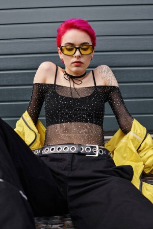 jeune femme aux cheveux roses et tatouage posant en lunettes de soleil jaunes et streetwear tendance dans la rue
