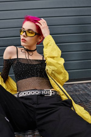 Foto de Mujer joven tatuada y de pelo rosa con gafas de sol amarillas y atuendo moderno posando en la calle urbana - Imagen libre de derechos