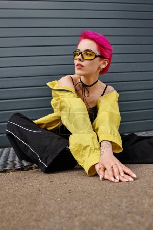 Foto de Mujer joven tatuada y de pelo rosa en gafas de sol amarillas y elegante atuendo posando en la calle urbana - Imagen libre de derechos