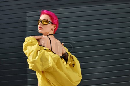rosafarbene junge Frau mit gelber Sonnenbrille und modischem Outfit posiert und blickt in die Kamera