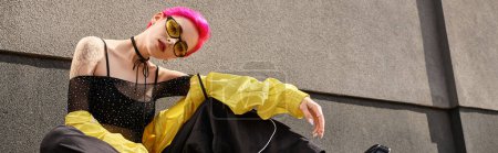 Foto de Longitud completa, mujer de pelo rosa en gafas de sol amarillas y traje de moda posando y mirando a la cámara - Imagen libre de derechos