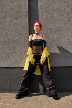Téléchargez les photos : Femme aux cheveux roses en lunettes de soleil jaunes et tenue tendance posant près du mur gris sur la rue urbaine - en image libre de droit