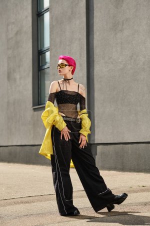 Foto de Mujer de pelo rosa en gafas de sol amarillas y traje de moda posando cerca del edificio en la calle urbana - Imagen libre de derechos