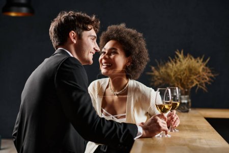 Téléchargez les photos : Joyeuse dame afro-américaine tenant verre de vin et regardant l'homme pendant la date sur la Saint-Valentin - en image libre de droit