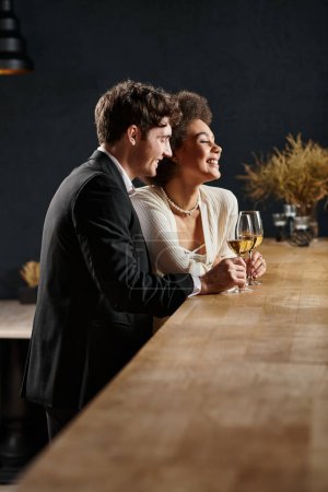 glückliches multikulturelles Paar im Abendkleid mit Gläsern Weißwein und lächelnd beim Date