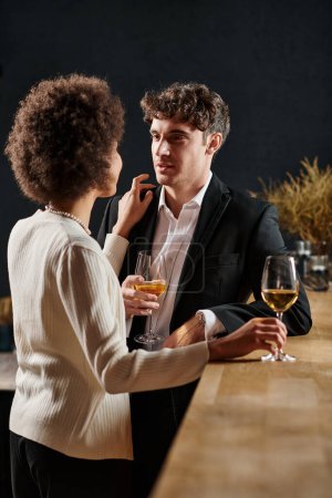 handsome man looking at african american girlfriend during date on valentines day, wine glasses