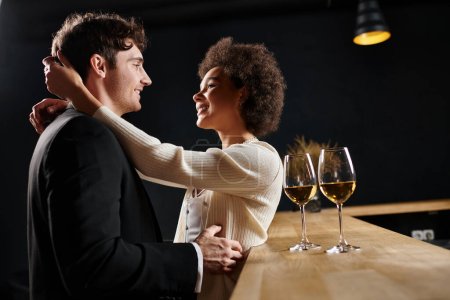 happy interracial couple embracing near glasses of wine on bar counter during date on Valentines Day