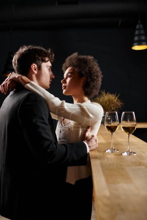 cariñosa pareja interracial abrazando cerca de copas de vino en el mostrador del bar durante la fecha en el restaurante
