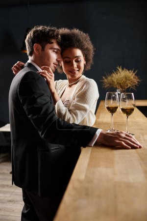 cariñosa pareja interracial abrazando y mirando copas de vino en el mostrador del bar durante la fecha