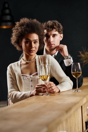attraktives multikulturelles Paar blickt in die Kamera, während es mit Weingläsern in der Nähe der Theke steht