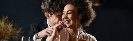 happy multicultural couple laughing and sitting at bar counter with wine glasses during date, banner