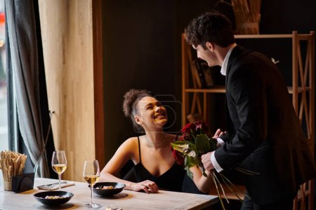 Herr im Anzug hält Rosen in der Nähe einer glücklichen afrikanisch-amerikanischen Frau in einem Restaurant am Valentinstag