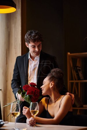 schöner Mann im Anzug mit Rosen in der Nähe einer glücklichen afrikanisch-amerikanischen Frau in einem Restaurant am Valentinstag