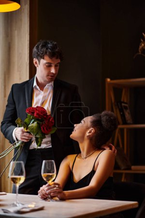 Photo for Happy man in suit holding roses near young african american woman in restaurant on Valentines Day - Royalty Free Image
