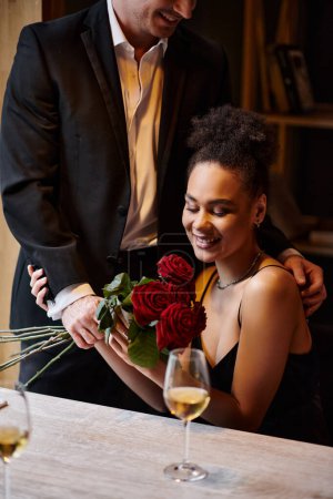 hombre de traje sosteniendo rosas cerca de la mujer afroamericana feliz en el restaurante en el día de San Valentín