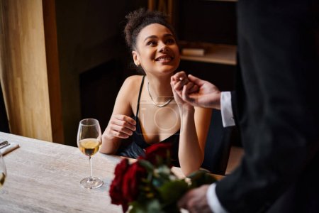 Mann im Anzug hält Hand einer glücklichen afrikanisch-amerikanischen Frau in Restaurant am Valentinstag