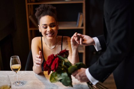 Mann im Anzug hält Hand einer fröhlichen afrikanisch-amerikanischen Frau in Restaurant am Valentinstag