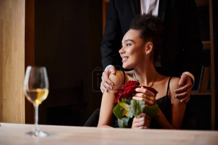 pleased african american woman holding red roses near man in suit standing behind her during date