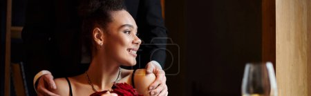 Photo for Pleased african american woman holding red roses near man standing behind her during date, banner - Royalty Free Image