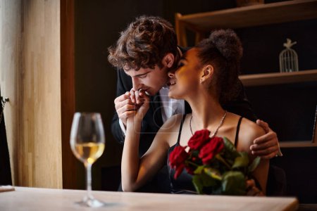 Photo for Gentleman kissing hand of happy african american woman holding roses during date on Valentines day - Royalty Free Image
