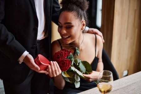 caballero regalando caja en forma de corazón a la mujer afroamericana feliz con rosas en el día de San Valentín