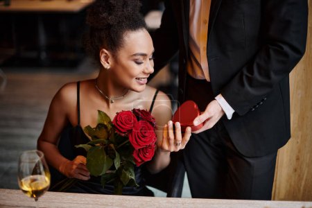 caballero regalando caja en forma de corazón a la mujer afroamericana feliz con rosas rojas en el día de San Valentín