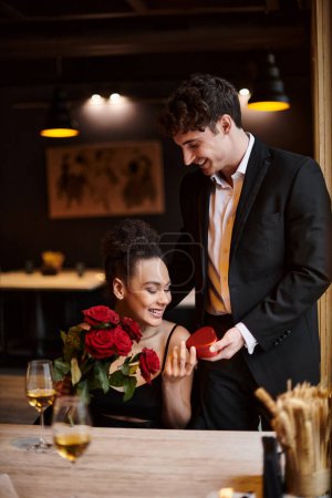 caballero regalando caja en forma de corazón a la mujer afroamericana feliz con rosas rojas el 14 de febrero