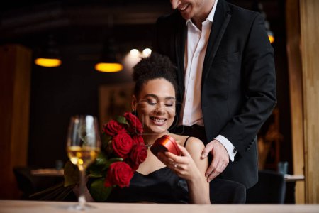 feliz hombre regalando caja en forma de corazón a la alegre mujer afroamericana con rosas rojas el 14 de febrero