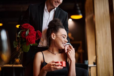 homme tenant des roses rouges près heureuse femme afro-américaine avec boîte en forme de coeur sur la Saint-Valentin