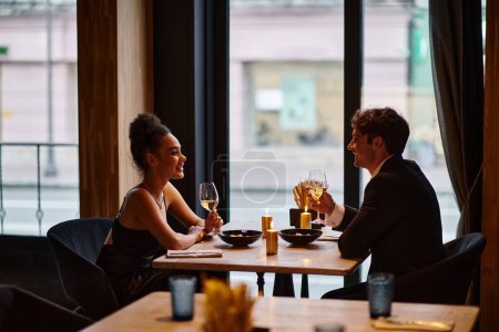 Photo for Happy interracial couple in elegant attire holding glasses with wine during date in restaurant - Royalty Free Image