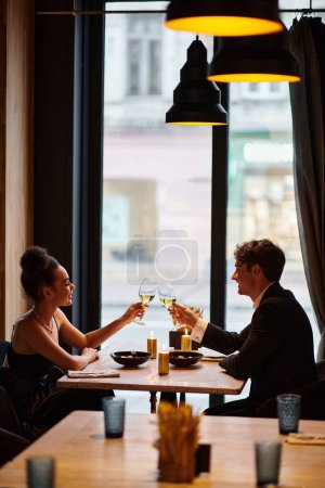 happy interracial couple in elegant attire toasting glasses with wine during date in restaurant