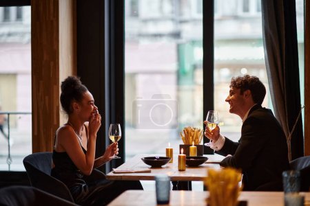 glückliches gemischtrassiges Paar in eleganter Kleidung mit Weingläsern beim Date im Restaurant, lacht