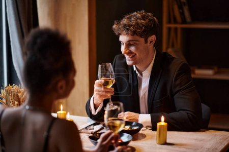 joyful man in elegant attire looking at glass of wine near woman during date in restaurant
