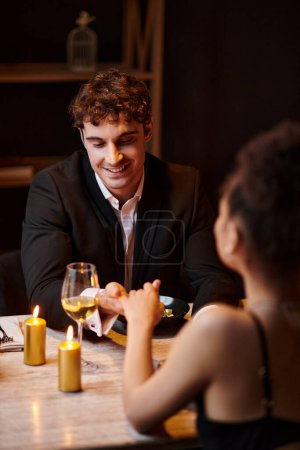 fröhlicher Mann in eleganter Kleidung, der bei einem Date im Restaurant auf die Hand seiner Freundin schaut, Valentinstag