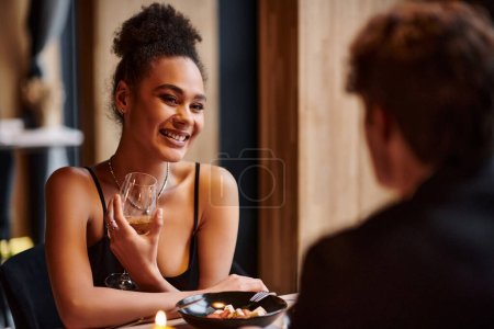 cheerful african american woman holding wine glass next to boyfriend during date on Valentines day