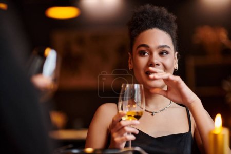 Photo for Young african american woman smiling and holding glass of wine during date on Valentines day - Royalty Free Image