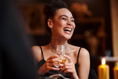 Photo for Cheerful african american woman laughing and holding glass of wine during date on Valentines day - Royalty Free Image