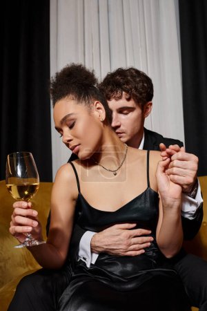 man hugging seductive african american woman holding glass of wine and sitting on velvet sofa