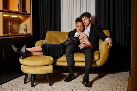Téléchargez les photos : Beau couple multiculturel en tenue de soirée noire posant ensemble tout en étant assis sur un canapé en velours - en image libre de droit