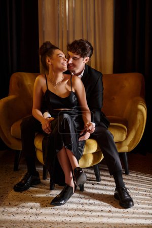 Téléchargez les photos : Bel homme en costume embrassant femme afro-américaine gaie en robe noire et assis sur le canapé - en image libre de droit