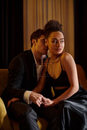 Téléchargez les photos : Bel homme en costume séduisant jolie femme afro-américaine en robe noire et assise sur le canapé - en image libre de droit