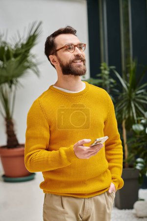 joyous handsome architect with glasses in yellow turtleneck posing with his phone in his office