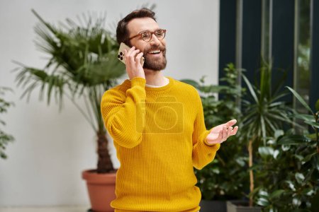 cheerful handsome architect with glasses in yellow turtleneck talking by his phone in his office