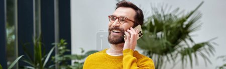 joyful handsome architect with glasses in yellow turtleneck talking by his phone in his office