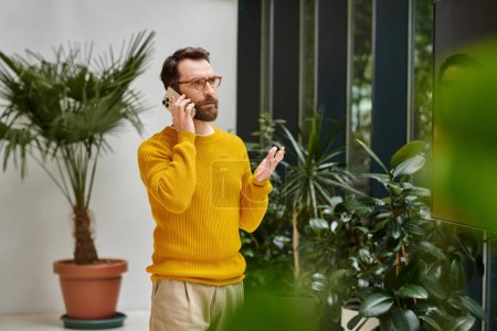 concentrated attractive bearded man in yellow turtleneck talking by his mobile phone in office