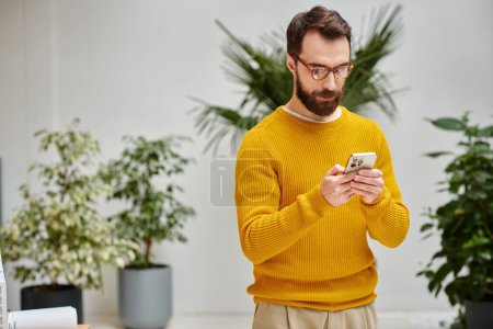 concentrated attractive bearded man in yellow turtleneck holding his mobile phone in office